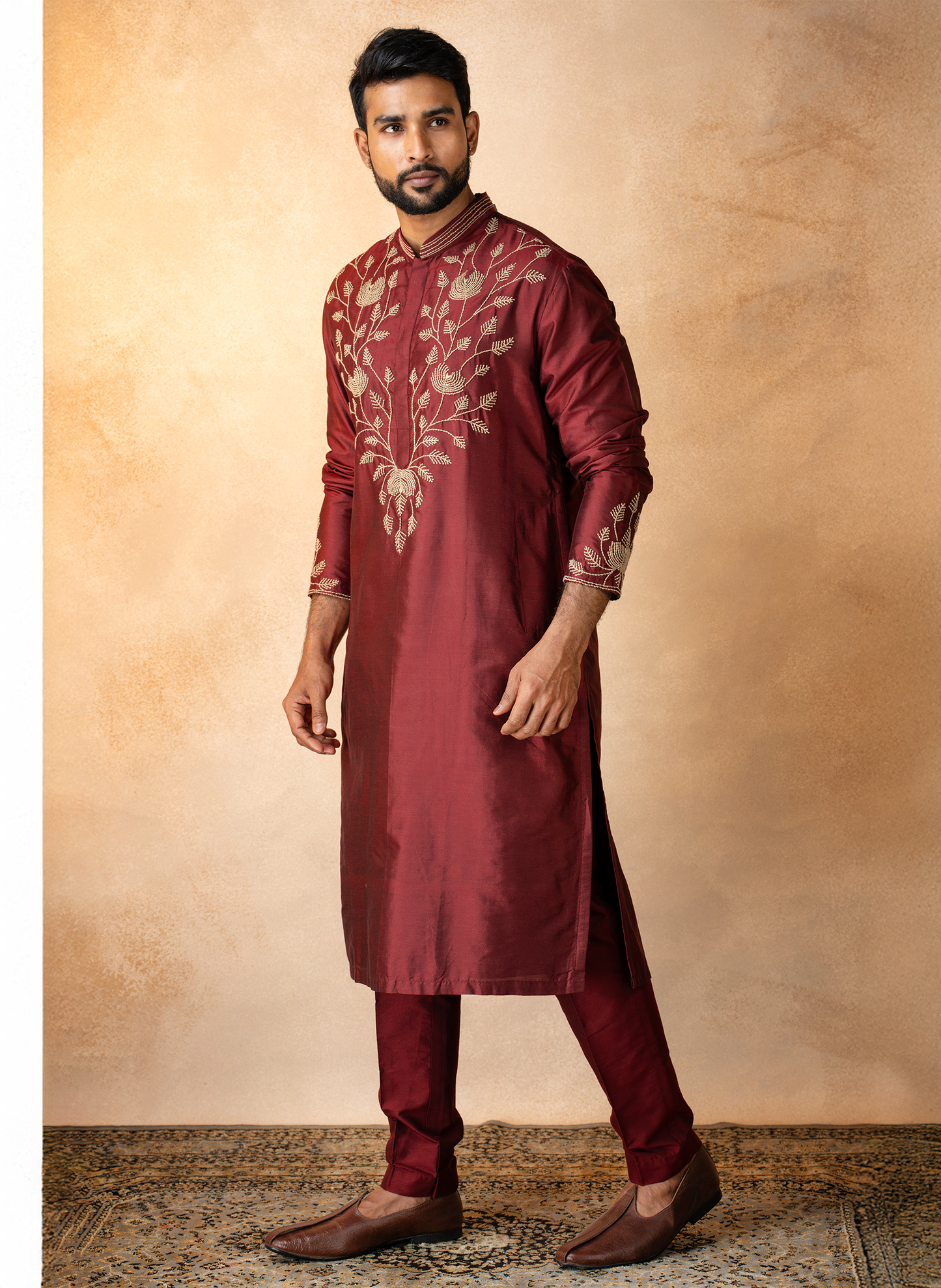 Man wearing a maroon embroidered kurta with pants against a beige wall.