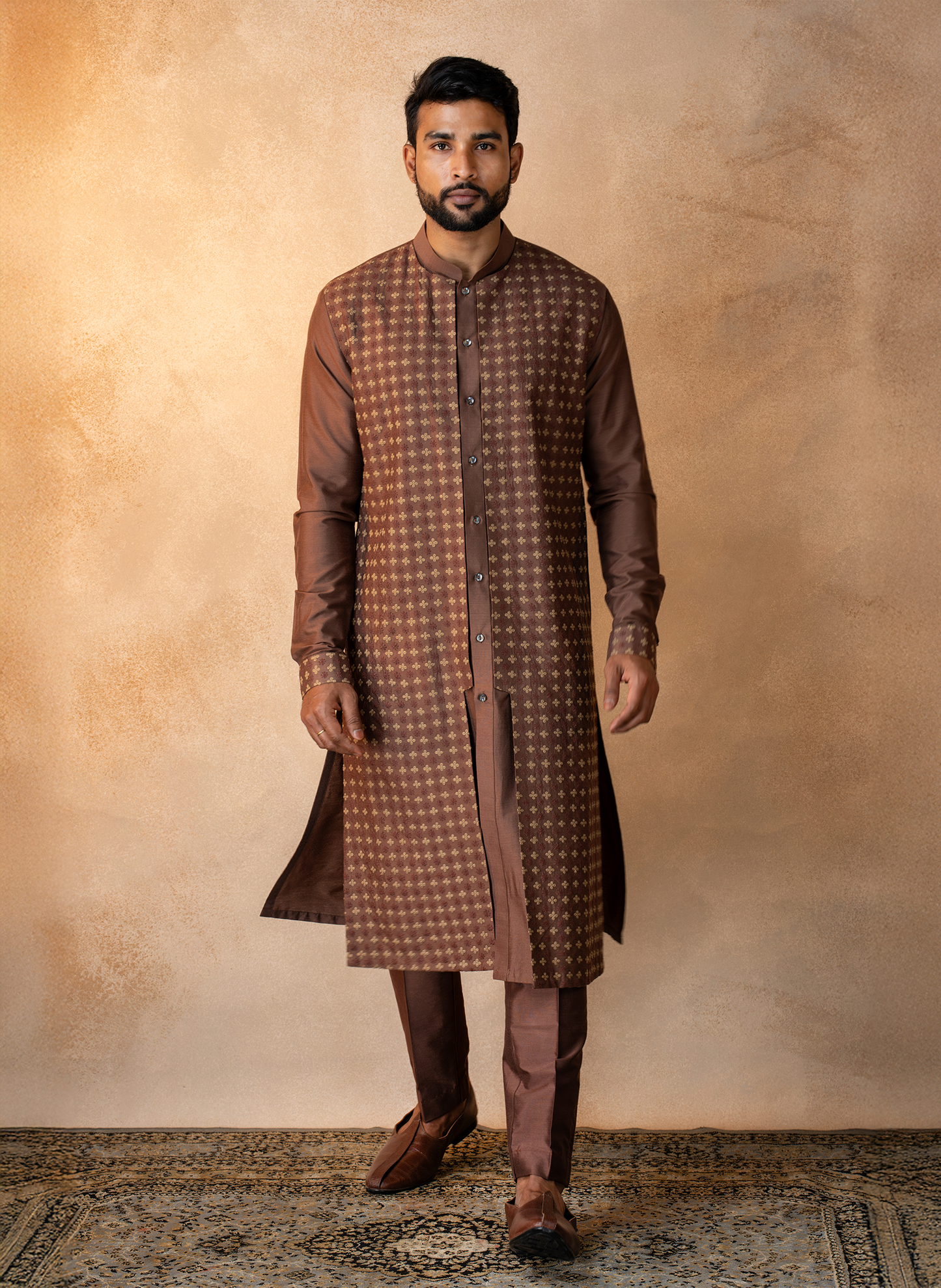 Man wearing a brown embroidered kurta with pants against a textured beige wall.